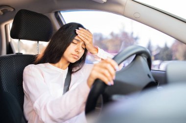 Arabanın içinde eli alnında yorgun bir Hintli kadın, hasta ya da bunalmış görünüyor. Araba sürerken zihinsel yorgunluğu, stresi veya fiziksel rahatsızlığı yansıtır.