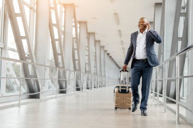 Siyah işadamı telefonda konuşurken havaalanında yürüyor, bavulunu çekiyor. Kendinden emin ve gülümseyen, şık giyinmiş ve iş seyahatine hazır, kopyalama alanı