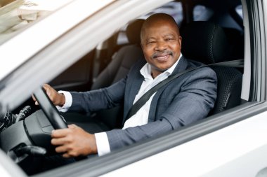 Siyahi işadamı güvenle arabanın koltuğunda oturuyor, camdan gülümsüyor. Takım elbise giymiş, yolculuğuna başlamadan önce sakin ve profesyonel görünüyor. Yakın plan.