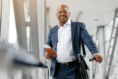 Profesyonel siyah işadamı havaalanında gülümserken pasaport ve bavul tutuyor. Şık ve kendinden emin, rahat ve iyimser bir sonraki durak için hazır, boşluğu kopyala