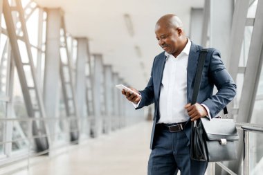 İş adamı havaalanında hafif bir gülümsemeyle akıllı telefon ekranı okuyor. Afrika kökenli Amerikalı erkek çantalı ve takım elbiseli. Odaklanmış ve dijital iletişimle meşgul görünüyor.