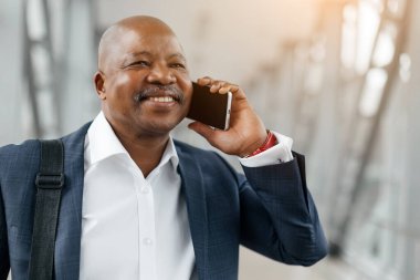 Telefonda gülümseyen siyah iş adamının yakın plan portresi. İfadesi rahat ve meşgul, takım elbise giyiyor ve açık bir havaalanı terminalinde duruyor.