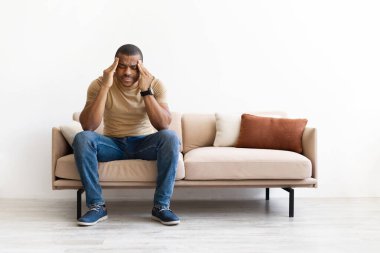 Kanepede oturan genç siyahi adam, elleri şakaklarda, stres ya da baş ağrısı çekiyor. Rahat ev ortamında gelişigüzel tükenme veya zihinsel yorgunluk ifadesi, boşluğu kopyala