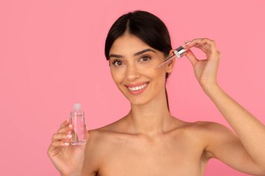 Gülümseyen genç bir kadın yüz serumunu düşürücüyle uyguluyor. Taze cilt bakımı, hidrasyon ve güzellik tedavisini minimalist pembe arka planda, yakın plan çekimlerinde teşvik eder.