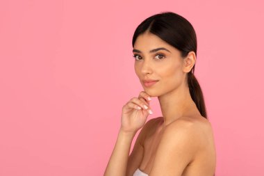 Zarif, genç bir kadın, eli çenesine yakın poz veriyor. Pembe stüdyo arka planında açık ten ve doğal zarafeti gösteren yumuşak güzellik portresi, kopyalama alanı