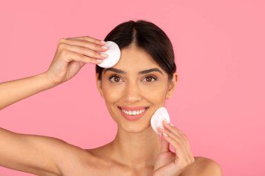 Yüzünde ve alnında pamuk pedlerle gülümseyen genç bir kadın. Taze ve eğlenceli cilt temizleme anı. Pembe arka planda doğal güzellik, kopyalama alanı.