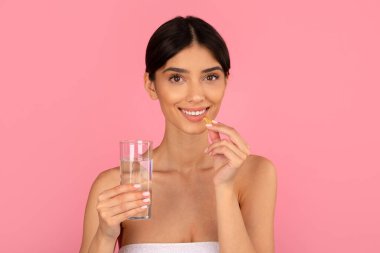 Gülümseyen kadın bir bardak suyla kapsül takviyesi alıyor. Sağlık, beslenme ve güzelliği içten dışa canlı pembe arka planda, kopyalama alanı