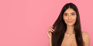 Güzel, uzun saçlarını nazik bir gülümsemeyle gösteren mutlu genç bir kadın. Sağlıklı saç ve güzelliği yumuşak pastel pembe stüdyo arka planında tanıtmak, fotokopi alanı olan panorama