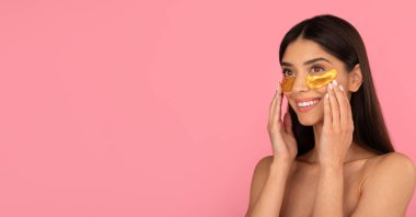 Gülümseyen genç bir kadın göz altı bantlarına dokunuyor. Cilt bakımı ve yaşlanma karşıtı güzellik ayini. Pembe arka planda taze bir görünüm. Kopya alanı olan bir manzara.