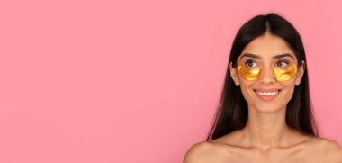 Altın rengi göz bandı takan ve gülümseyen genç bir kadın. Kabarıklık, ışıltı ve nemlilik için pembe arka planda güzellik ve cilt bakımı, kopyalama alanı olan panorama