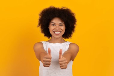 Kendinden emin, gülümseyen siyahi kadın onay ve pozitiflik gösteriyor. Canlı sarı arkaplan üzerine stüdyo çekimi, destek ve başarı ifade ediyor