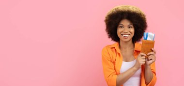 Hasır şapkalı, elinde seyahat biletiyle gülümseyen siyah bir kadın. Turizm ve yaz kaçamağı için stüdyo resmi, pembe arka planda izole edilmiş, kopya alanı olan panorama