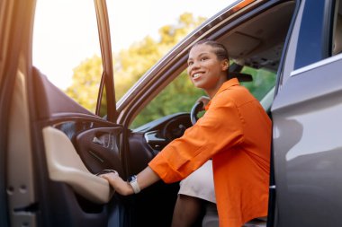 Gülümseyen genç siyahi kadın arabadan çıkıyor, kapıyı tutuyor ve başka tarafa bakıyor. Pozitif titreşim, özgürlük ve beklenti hareket halinde, yakın çekim