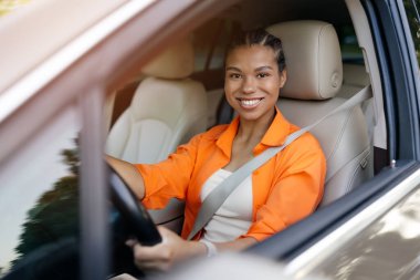 Turuncu elbiseli, kendinden emin genç siyahi bir kadın pencereden gülümsüyor. Güvenli, odaklanmış ve direksiyonun arkasında şık, yakın plan.