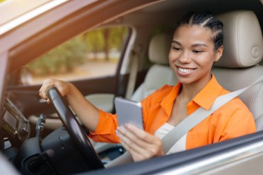 Turuncu bluzlu genç siyah kadın arabada otururken akıllı telefonunu kontrol ediyor. Sakin ve neşeli, dijital hayatı günlük işe gidip gelme ile harmanlayan, araba kullanırken birden fazla iş yapan