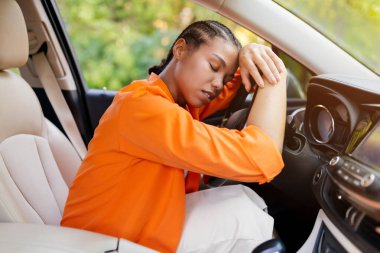 Yorgun, genç, siyah bir kadın arabanın içinde direksiyonda dinleniyor. Gözler kapalı, açıkça boğulmuş ya da yorgun, muhtemelen uzun bir yolculuktan ya da stresli bir günden sonra, yakın çekim