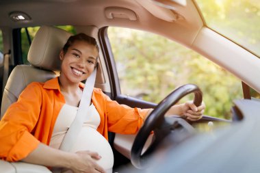 Hamile siyah kadın araba kullanırken gülümsüyor, bir eli direksiyonda, diğeri de karnında. Sıcak, neşeli bir an. Hamileliğin sonlarında kendine güvenini ve sakinliğini ifade ediyor. Yakın plan.