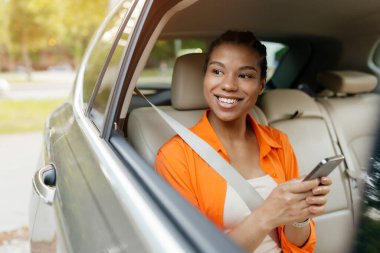 Neşeli siyah kadın arabanın arka koltuğunda oturmuş elinde akıllı telefonuyla pencereden dışarı bakıyor. Bağlı ve mevcut, sürüşün tadını çıkarıyor, yakın plan