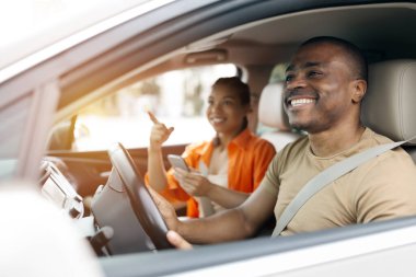 Genç siyah çift birlikte araba kullanıyor ve akıllı telefon kullanıyor, sürüş sırasında gülüyor ve gülümsüyor, kadınlar telefonda erkeklere yol tarifi konusunda hevesle yardım ediyor, kapatıyor.