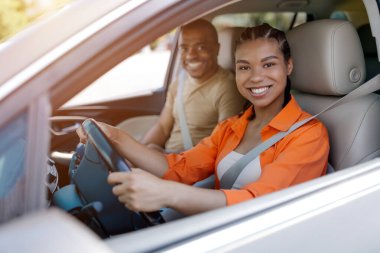 Araba kullanırken direksiyonda gülümseyen gururlu genç siyahi kadın şoför. Partneri destekle gülümsüyor, birlikte yolun tadını çıkarıyor. Kapanıyoruz.