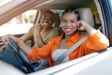 Gülümseyen genç siyahi kadın, erkek arkadaşıyla güvenli bir şekilde araba kullanıyor, kolunu dinlendiriyor ve sürüşün tadını çıkarırken, ortağı da onu sıcak bir ifadeyle destekliyor..