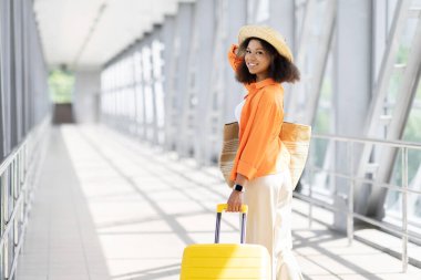 Sarı bavulla yürüyen genç siyahi kadın yolcuların arka görüntüsü. Havaalanında kendine gülerek arkasına bakıyor, şık bir kaçış için giyinmiş, mekanı kopyalıyor.