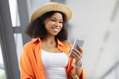Havaalanındaki telefon ekranına gülümseyen siyahi kadın, mesajın keyfini çıkaran hasır şapkalı kadın ya da seyahat uygulaması güncellemesi. Rahat ve neşeli bir ortam, kopyalama alanı
