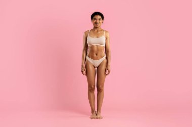 Bej iç çamaşırlı Afro-Amerikalı genç bir kadın, pembe bir arka planda güvenle duruyor, doğal bir şekilde gülümsüyor, atletik fizikli, kopyalama alanı