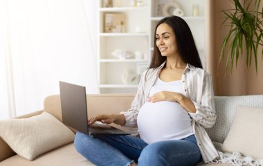 Kanepede oturan hamile bir kadın dizüstü bilgisayar kullanıyor, sakince gülümsüyor. Uzaktan çalışmayı, çevrimiçi eğitimi veya hamilelik süresince dijital iletişimi temsil eder, kopyalama alanı