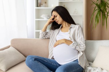 Hamile kadın elleriyle alnına dokunuyor. Evde baş ağrısı, stres ya da yorgunluk belirtileri gösteriyor. Hamileliğin hormonal ve duygusal bedelini yansıtan gerçekçi bir an.