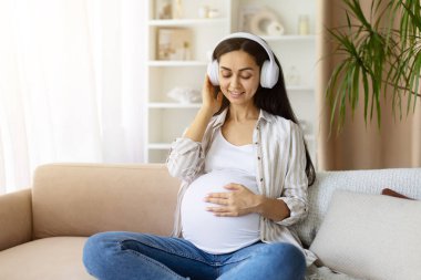 Hamile bir kadın kablosuz kulaklıkla müzikten hoşlanıyor, karnına hafifçe dokunuyor. Hamilelik sırasındaki ses yoluyla sakin ve neşeli bir bağ ve rahatlama anı