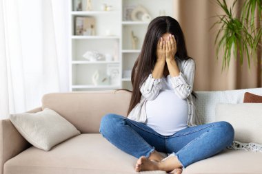 Hamile bir kadın kanepede bacak bacak üstüne atarak oturuyor, elleriyle yüzünü kapatıyor, evde ağlıyor endişe, korku ya da bunaltıcı duygular ifade ediyor. Hamilelik ruh hali değişikliklerinin dürüst tasviri