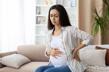 Hamile bir kadın koltukta oturup karnını ve yanını tutarken sırt ağrısı ve yorgunluk çekiyor. Hamilelikteki fiziksel rahatsızlığın gerçekçi görünümü, yakın plan.