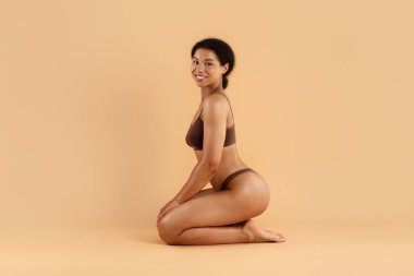 Kahverengi iç çamaşırlarıyla bej arka planda oturan gülümseyen kadın. Yumuşak ışıklandırma ve kendine güvenen pozlar refah, güzellik ve konfor kavramları için.