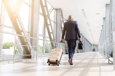 Modern havaalanı terminalinde bavulla yürüyen resmi takım elbiseli kendine güvenen siyah iş adamı. Seyahat, hareketlilik, küresel iş ve kurumsal yaşam tarzı, dikiz açısı sembolü