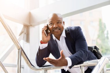 Havaalanında korkuluklara yaslanırken akıllı telefondan konuşan Afro-Amerikalı iş adamı. Profesyonel seyahat yaşam tarzı, iletişim ve kurumsal bağlantı