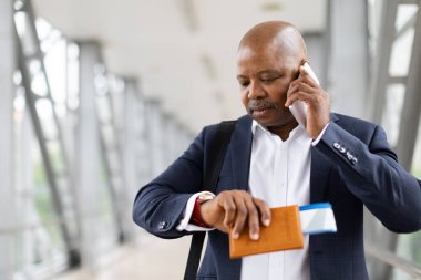 Akıllı telefonla konuşurken ve havaalanı terminalinde pasaportu elinde tutarken Afrika kökenli Amerikalı bir işadamı saati kontrol ediyor. İş seyahati ve dakiklik kavramı, yakın plan.