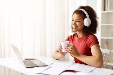 Evde dizüstü bilgisayarla çalışırken kulaklıklı Afro-Amerikalı bir kadın kahvenin tadını çıkarıyor. Serbest yaşam tarzı kavramı, çevrimiçi eğitim veya uzak ofis verimliliği