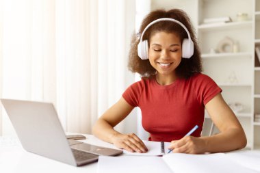 İnternette okurken masasında notlar yazan kulaklıklı, gülümseyen Afrikalı Amerikalı kadın. Uzaktan öğrenme, serbest çalışma ya da dizüstü bilgisayarlı ev işi yaşam tarzı