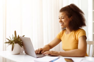 Bilgisayarda bitkilerle yazı yazan gülümseyen Afrikalı Amerikalı kadın. Modern ev ofisinde uzaktan çalışma, çevrimiçi öğrenme ve serbest iş hayatı