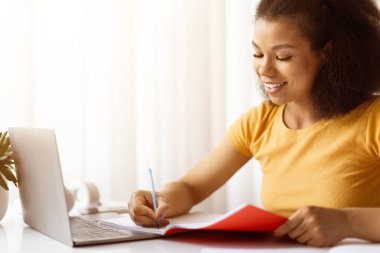 Gülümseyen Afrikalı Amerikalı kadın dizüstü bilgisayar masasında kırmızı not defterine notlar yazıyor. Uzaktan öğrenme, serbest çalışma projeleri ya da evdeki modern online öğrenci yaşam tarzı