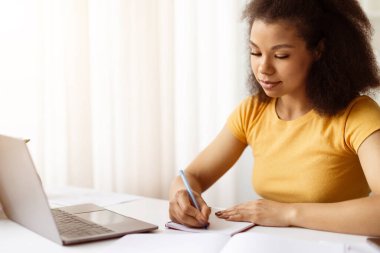 Laptopta çalışırken not defterine notlar yazan odaklanmış Afrikalı Amerikalı kadın. Uzaktan öğrenme, serbest çalışma projeleri veya evde profesyonel çevrimiçi eğitim