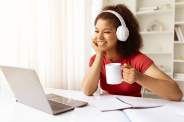 Kulaklıklı Afro-Amerikalı kadın laptopa bakarken kahve içiyor. Modern bir ofis ortamında uzaktan çalışma, çevrimiçi öğrenme ve serbest yaşam tarzı