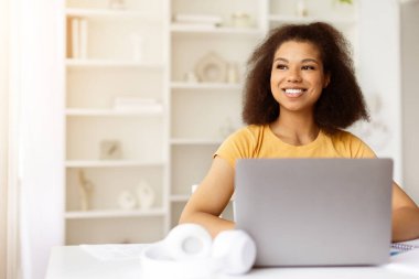 Hayalperest Afrikalı Amerikalı kadın dizüstü bilgisayar başında çalışırken gülümseyip başka tarafa bakıyor. Pozitif ruh hali, serbest kariyer ya da ofis masasında online eğitim yaşam tarzı