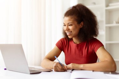 Gülümseyen Afrikalı Amerikalı kadın dizüstü bilgisayarla notlar yazıyor. Uzaktan öğrenim, serbest çalışma projeleri ve ev ofisinde modern online eğitim yaşam tarzı