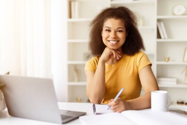 Dizüstü bilgisayarla çalışırken kameraya gülümseyen kendinden emin Afrikalı Amerikalı kadın. Çevrimiçi öğrenim, serbest çalışma veya modern ofis yaşam tarzı kavramı