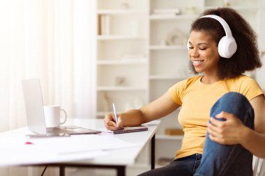 Laptopta çalışırken kulaklıklı, gülümseyen Afrikalı Amerikalı kadın notlar yazıyor. Çevrimiçi eğitim, serbest kariyer veya modern dijital yaşam tarzı kavramı