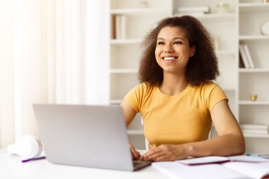 Ofiste bilgisayarla gülümseyen Afrikalı Amerikalı kadın. Uzaktan çalışma kavramı, serbest çalışma projeleri, çevrimiçi ve çağdaş dijital yaşam tarzı eğitimi