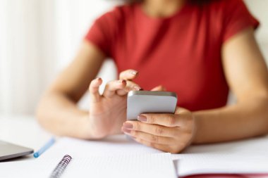 Afrika kökenli Amerikalı bir kadının masa başında elinde defterlerle akıllı telefon kullanması. Dijital iletişim, çevrimiçi çalışma, uzaktan çalışma ve ev kavramında serbest yaşam tarzı
