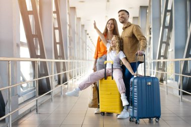 Üç kişilik beyaz bir aile havaalanında, bavulları fotokopi odasına bakıyor, kızı bavulun üzerinde oturuyor, ailesi yanında gülümsüyor, neşeli bir yaşam tarzı, birlik ve keyifli bir tatil seyahati sergiliyor.